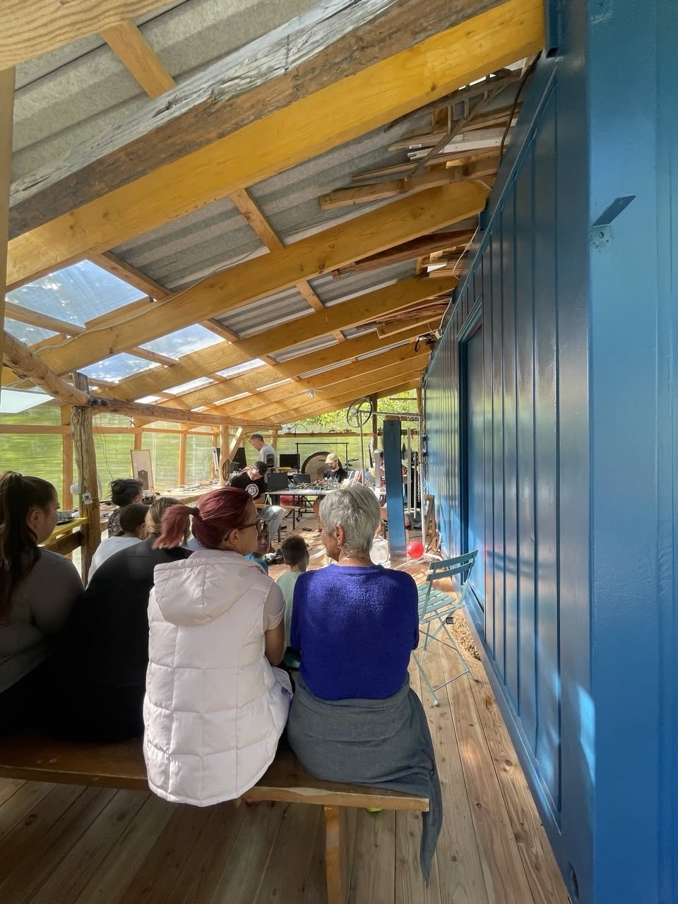 Neighbours seated on a bench in the greenhouse beside the KontejnerLab, watching and joining the improvisation session